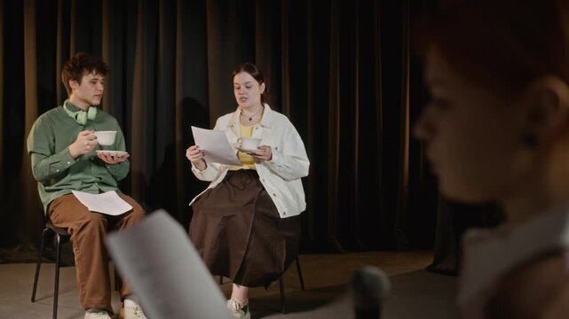 Rack focus shot of Caucasian teenage girl holding microphone and reading script with other teenagers rehearsing scene with teacups on stage. For promoting school drama clubs and youth arts education