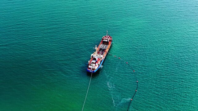 Dredging vessel with trailing boom on turquoise sea. Aerial view of blue and red ship navigating open water, orange marker buoy and cable extending behind stern.