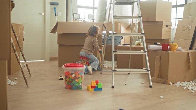 Little blonde girl riding a wooden trike among packed cardboard boxes and metal ladder in a building; playful curiosity.