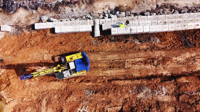 Workers installing concrete seawall blocks on eroded coast. Overhead shot of yellow excavator and worker in hi-vis vest placing precast block row beside rocky beach and calm sea.