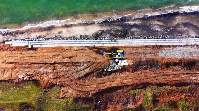 Aerial view of seawall construction with excavator and block placement on Black Sea shore. Drone shot of yellow excavator, concrete block row, rock riprap and turquoise wave-washed beach in daylight.