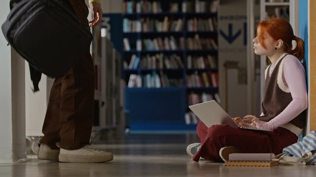 Side view of Caucasian teenage girl sitting on library floor and typing on laptop, joined by Caucasian teenage boy sitting down to discuss school project. Footage for high school education campaigns
