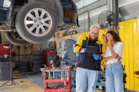 A mechanic is inspecting a car engine with a tool while explaining the issue to a customer in an auto repair shop