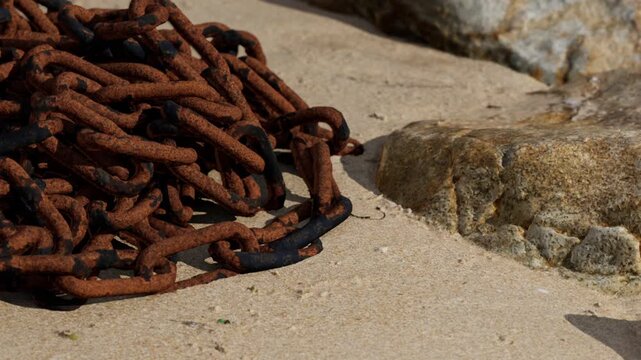 A pile of rusty metal chains is lying on the sand, next to some rough stones. The sun is shining brightly, highlighting the texture of the sand and the chains.