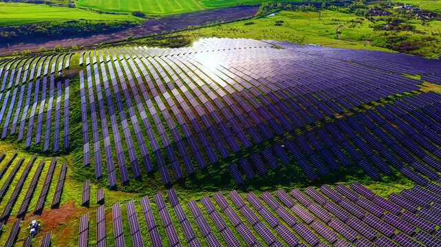 Massive solar power plant on a grassy hill. Elevated drone shot of numerous photovoltaic rows covering a rolling hillside with a bright central sun glint.