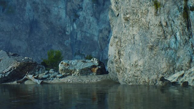 The scene shows large rocks beside calm water. Small plants grow near the waters edge. The surrounding area has a rocky cliff and clear reflections on the water.