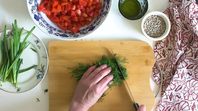 A woman prepares to make a salad by cutting fresh dill on a wooden board. The kitchen has vegetables like tomatoes and greens. Ingredients show a healthy meal preparation