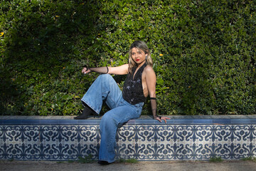 Fototapeta premium Young woman, Hispanic, beautiful, blonde, wearing baggy jeans, a black top, and fishnet gloves; relaxed and calm, sitting on a bench soaking up the sun. Concept: beauty, fashion, trends.