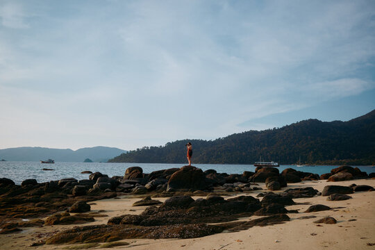 Person stands on rocks by water with boats in background