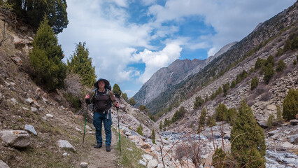 Fototapeta premium A man enjoys a captivating hike along a rugged trail in the Pamir Mountains of Kyrgyzstan. Towering peaks surround him, with lush greenery and a rushing river creating a beautiful backdrop.