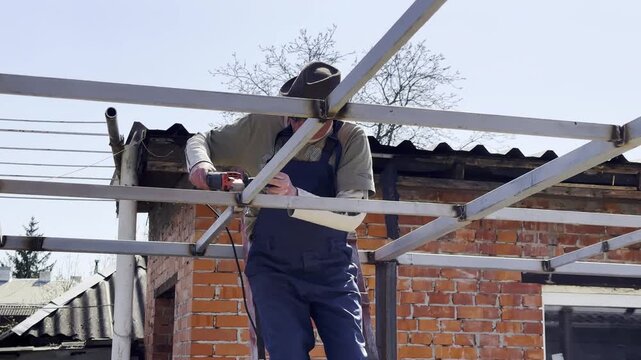 Adult worker polishing iron using electric grinder wheel on carport frame. Professional repairman grinding metal detail with circular saw at construction at sunny day. Male laborer working at the site