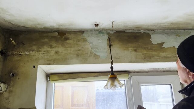 Male hands use putty knife to scrape and remove old plaster from an interior wall. Worker carefully cleans surface during renovation, preparing the wall for new finishing and repair work inside house