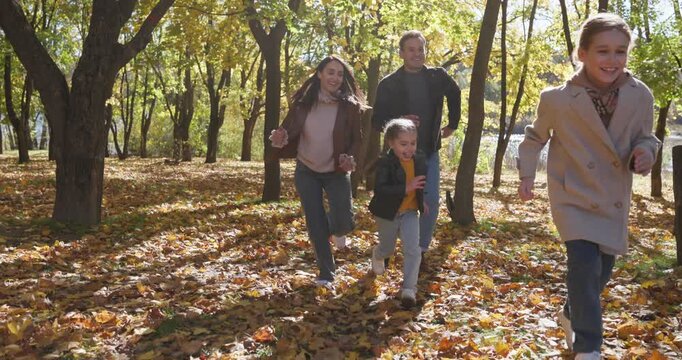 Happy family playing together on walk in autumn park. Parents and children running through forest laughing, mother and father chase and guide kids across golden leaves, enjoy time outdoors and nature