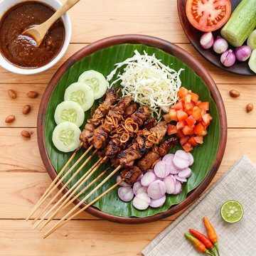Indonesian Traditional Sate Kambing Mutton Satay on Clay Plate with Banana Leaf, Flat Lay View of Asian Grilled Meat Skewers with Peanut Sauce, Raw Shallots and Vegetables on Wooden Table