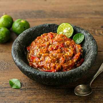 Indonesian Traditional Sambal Ulek Red Chili Paste in Stone Mortar, Three Quarter View of Asian Spicy Condiment with Lime Slice, Fresh Jeruk Limau and Antique Spoon on Rustic Wooden Table