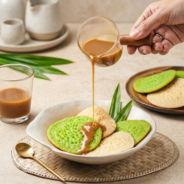 Hand Pouring Sweet Palm Sugar Sauce on Serabi, Indonesian Traditional Green Pandan and Coconut Pancakes, Drizzling Kinca Syrup on Rice Cake, Bright Cream Background Food Photography