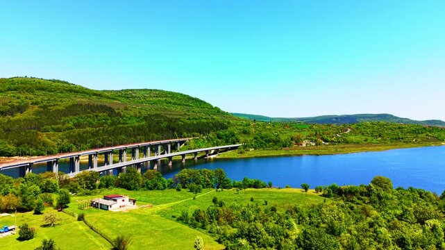 Railway bridge over reservoir, Bulgaria. Elevated angle of modern concrete viaduct spanning blue water surrounded by vibrant grassy landscape.