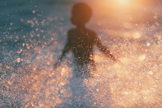 Child's silhouette splashing in water at sunset, with sunlight creating a sparkling effect on the surface and droplets captured in motion