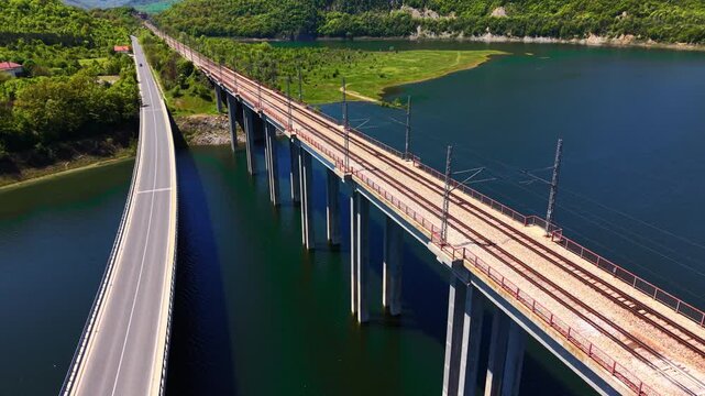 Railway bridge and road bridge, Bulgaria. High angle view of parallel infrastructure crossing a reservoir with green forest and hills.