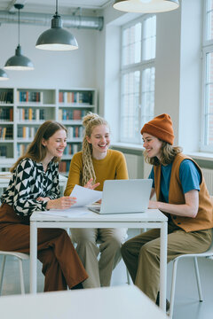 Vertical shot of three Caucasian young adult women collaborating at laptop, reviewing documents in library, representing teamwork and education. Useful for academic marketing or study services