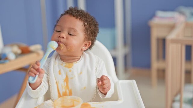 Toddler girl lifts spoon to mouth while self feeding in kitchen highchair with puree spilled on bib and tray; playful mealtime.