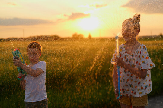 Two children have fun using water guns as the sun sets behind them in a green field. They laugh and splash water on each other in the evening glow