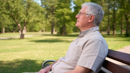 Fototapeta premium Senior man sitting on a park bench in a green landscape, wearing a light-colored shirt, enjoying a peaceful moment in nature with trees and grass surrounding him