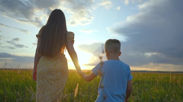 Mother and son walk in green field at sunset holding hands. Family moves through grass during summer evening. Mother and son enjoy nature outdoors together. Happy family goes forward in sunset field.