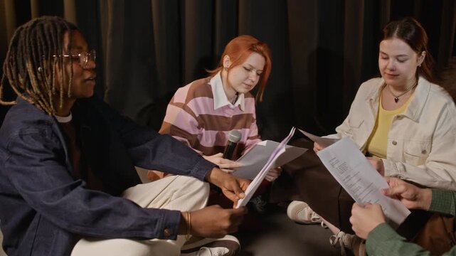 Panning shot of multiethnic teenagers sitting on stage floor, rehearsing play with scripts. Clip perfect for marketing drama schools, youth theater programs, or creative arts education