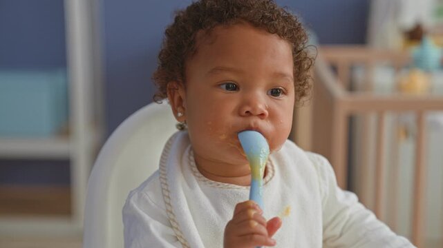 African american little girl eating with spoon in highchair, food smeared on bib and tongue out while self feeding inside building; playful curiosity.