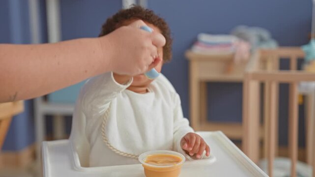 African american little girl eating baby food from a spoon held by an adult hand in a highchair in a building nursery, reaching for the spoon; tenderness bonding.