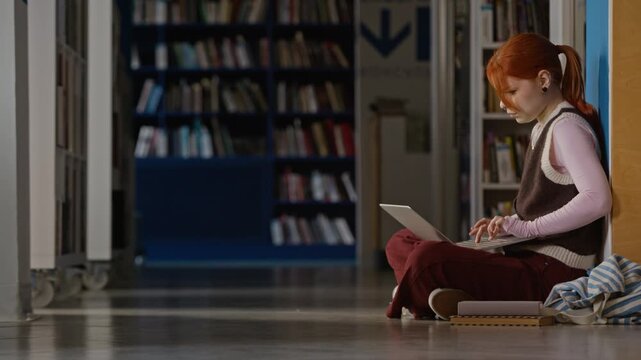 Side view of Caucasian teenage girl sitting on library floor, typing on laptop computer. Clip ideal for educational marketing, online learning platforms, and student lifestyle commercials