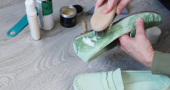 Woman hands brushing mint green suede loafer with active foam cleaner and wooden brush. Female person scrubbing casual nubuck moccasins using professional cleaning shampoo mousse during domestic
