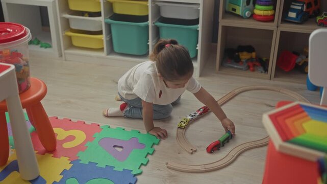 Girl kneeling, hand pushing toy car along wooden train track inside building playroom with foam mat and shelves; gentle concentration.