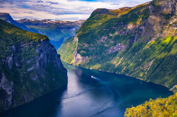 Panoramic view of Geirangerfjord with the Seven Sisters waterfall, steep green cliffs and a small boat on calm blue water in western Norway. © Nick Fox