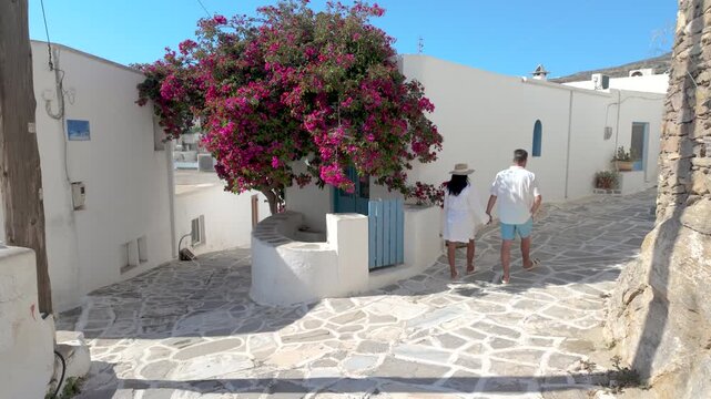 A diverse couple strolls through the picturesque streets of Naousa Paros, Greece, where colorful bougainvillea blooms adorn charming whitewashed buildings. Enjoy a leisurely walk under the sun.