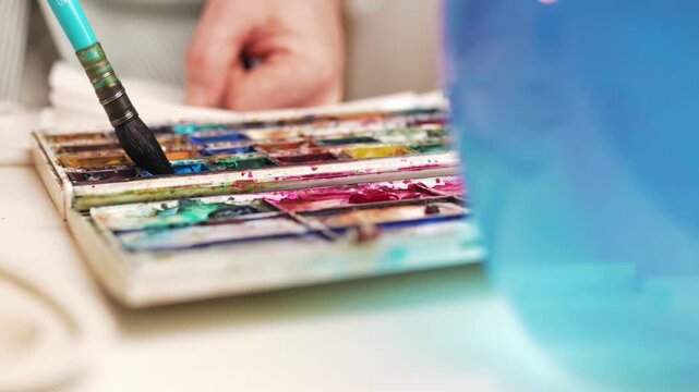 Closeup of artist hand dipping paintbrush into watercolor paint cuvette on palette