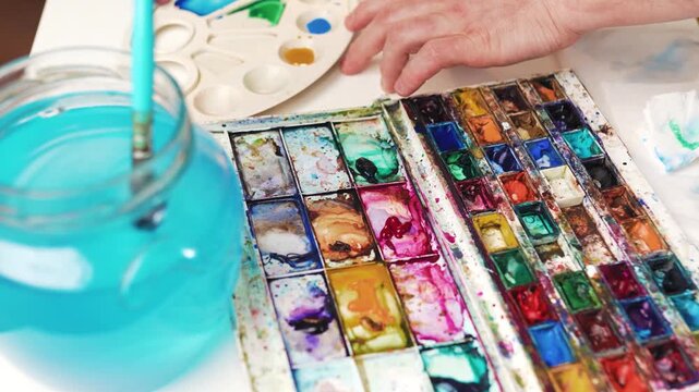 Paintbrush dipping into green watercolor cuvette with water jar beside