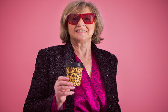 Mature woman with short gray hair wearing red sunglasses and a black sequined blazer holds a leopard print coffee cup against a pink background