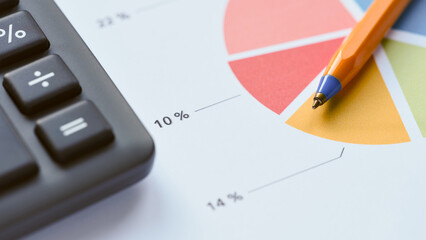 An orange pen lying on a colorful pie chart document next to a black office calculator, symbolizing financial planning and business analysis. Close up. © Stepan Popov