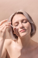 Naklejka premium Young woman with short blonde hair applying skincare serum to her face, showcasing a natural beauty routine in a soft, minimalistic setting with warm tones and gentle lighting