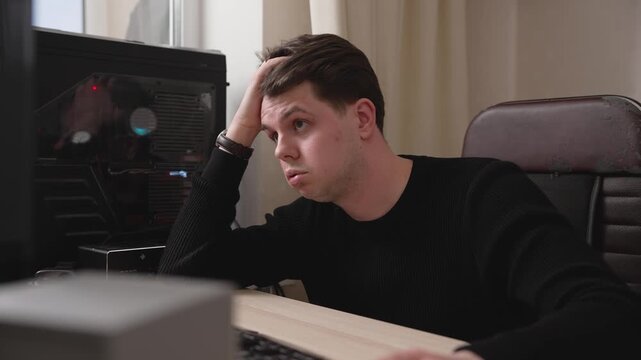 Young man in black sweater sitting at desk looking at computer screen with worried expression, hand on head, showing stress and frustration while working on PC
