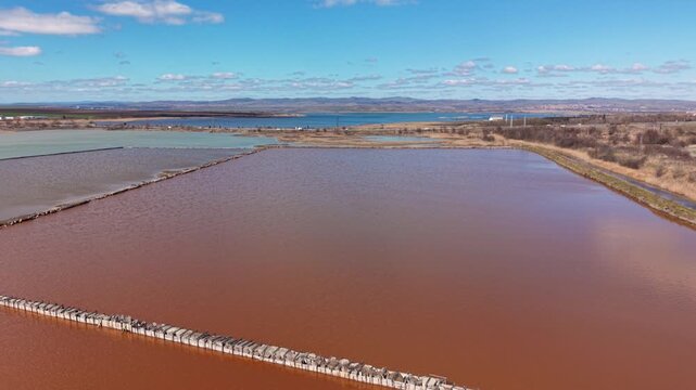 Aerial view of the pink salt pans of Lake Atanasovsko with reddish water and narrow embankments under a blue sky in Burgas, Bulgaria.