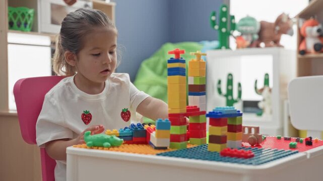Little girl stacking colorful plastic blocks into tall towers on a tabletop play table in a bright playroom with shelf toys, cactus decal and beanbag; quiet concentration.