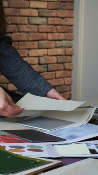 Hands sorting color swatches and paper samples on design table, adjusting printed sheets in creative workspace with brick wall backdrop and sample boards spread across surface., palette detail