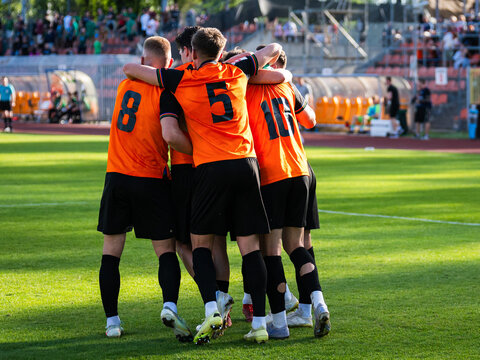 Soccer players in orange jerseys huddling and celebrating a goal during a football match on a sunny green pitch.