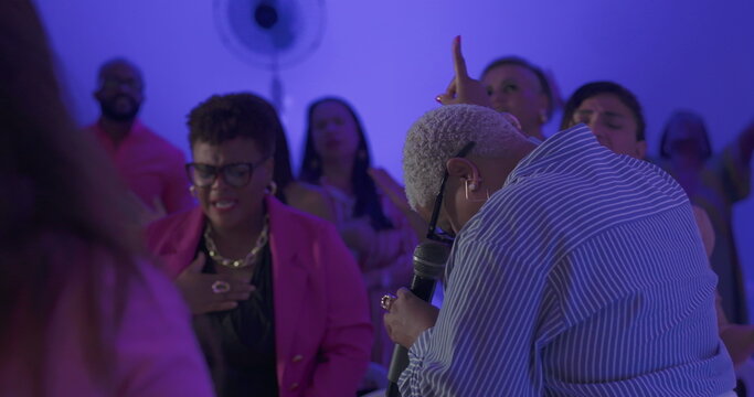 Preacher bows head in prayer and lifts microphone in passionate praise during evangelical church service as congregation worships with eyes closed and hands raised
