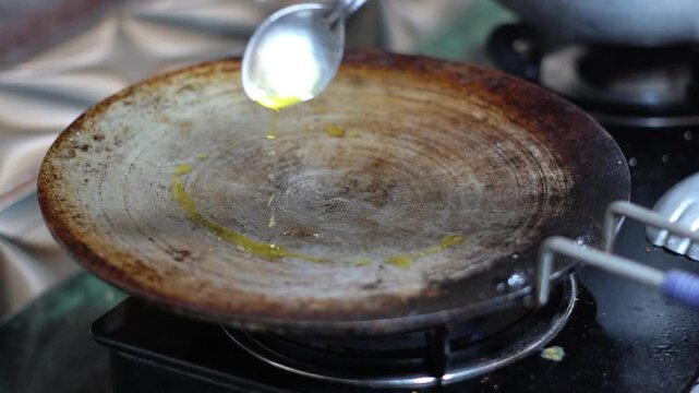 Pouring Cooking Oil from Spoon onto Hot Cast Iron Tawa Pan on Gas Stove. Drizzling Cooking Oil or Ghee on a Hot Flat Griddle Pan Close Up.