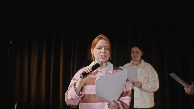 Medium shot of diverse teenagers rehearsing stage performance, passing microphone and reading scripts. Perfect for educational promos, drama school advertisements, and youth talent show concepts