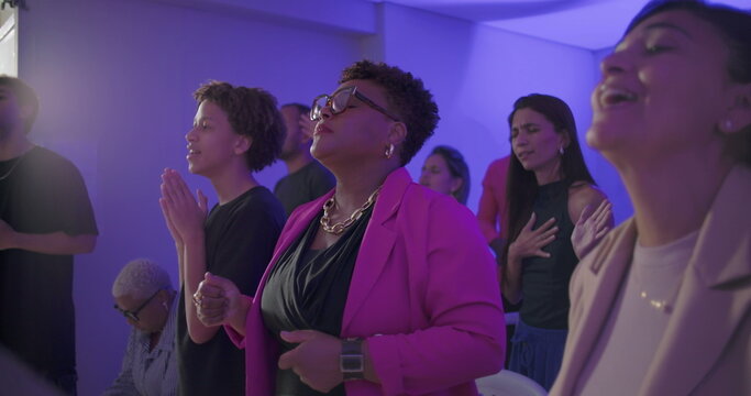 Woman raising arm in passionate prayer during evangelical worship service with group of diverse people standing and praying together in small indoor church